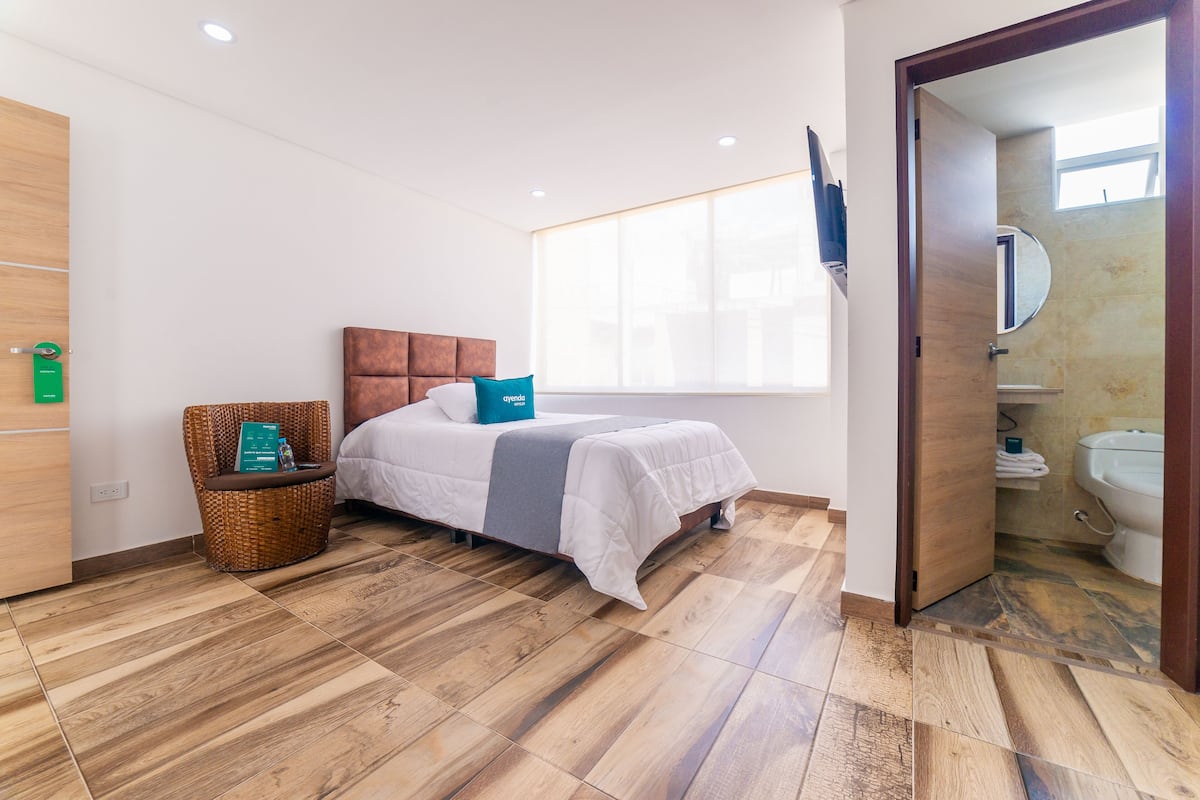 A modern hotel room with a wooden floor, a bed with white bedding, a wicker chair with a book, and a bathroom visible through an open door.