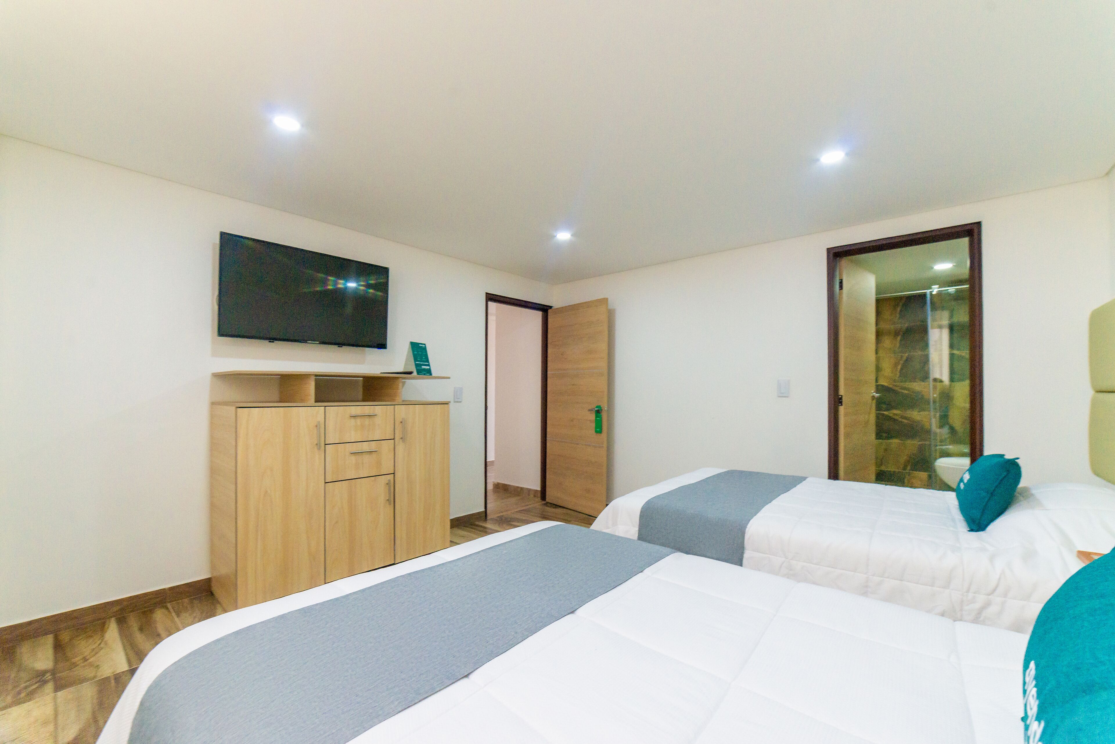 A hotel room with two beds, a TV mounted on the wall, a wooden cabinet, and a door leading to a bathroom.