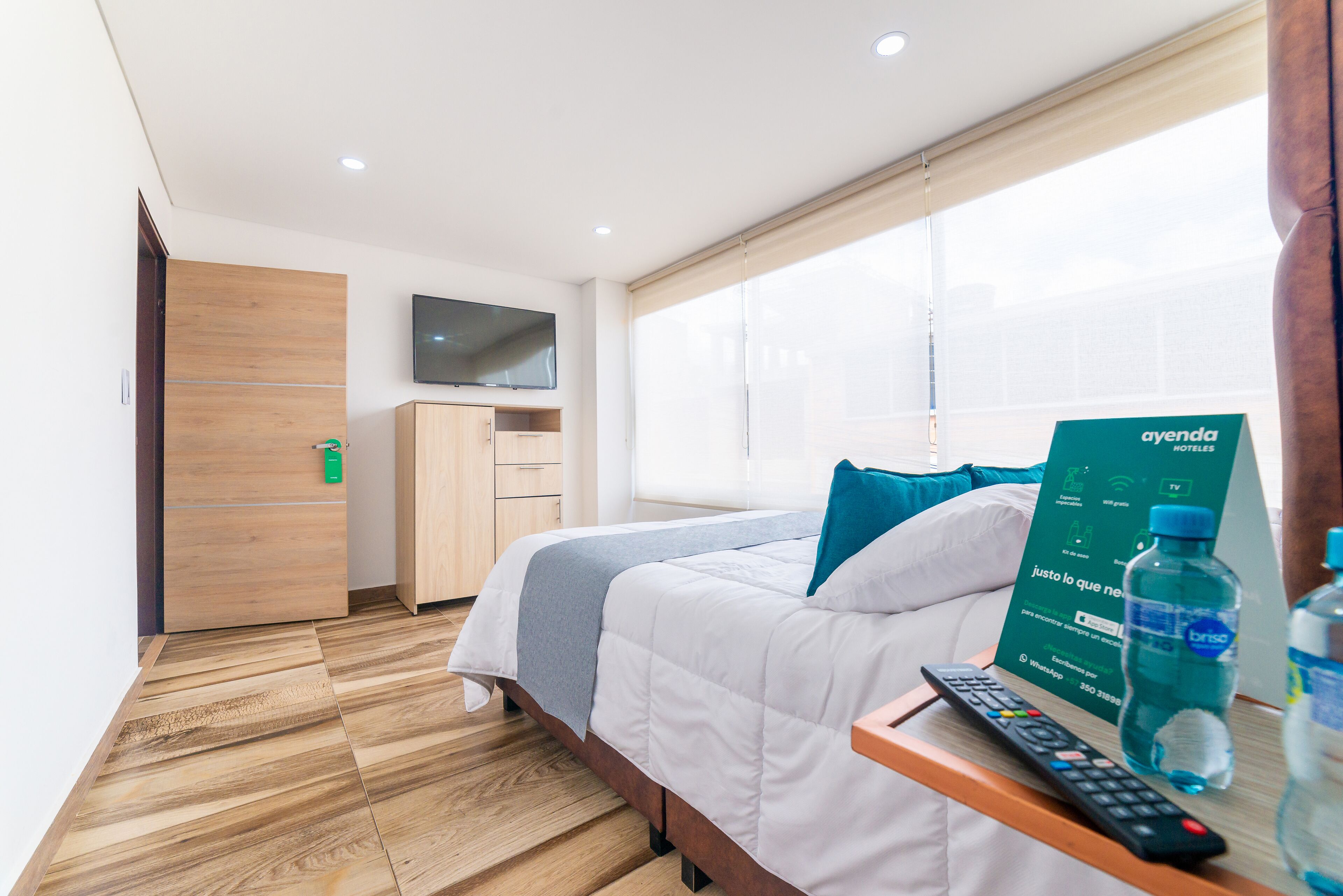 A hotel room with a bed, a TV mounted on the wall, a wooden wardrobe, and a bedside table with a remote control and water bottles.