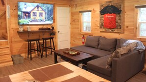 A cozy cabin interior with a television displaying a house, a sofa, a coffee table, and a small bar area. - Cabin in Wayne National Forest with ride in/out access to Baileys Trail System (Millfield)