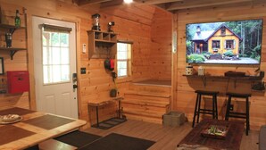 A wooden cabin interior with a dining area, kitchen, and a TV displaying a house outside. - Cabin in Wayne National Forest with ride in/out access to Baileys Trail System (Millfield)
