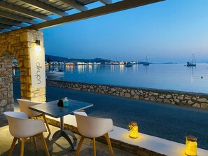 An outdoor seating area with stone pillars, a table, and chairs overlooking a calm body of water with boats.