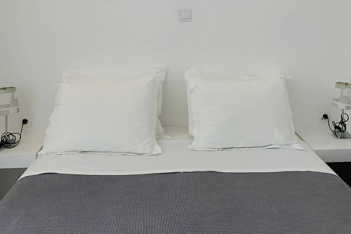 A bedroom with a bed, white pillows, a gray blanket, a wooden ceiling, and a nightstand with a lamp.