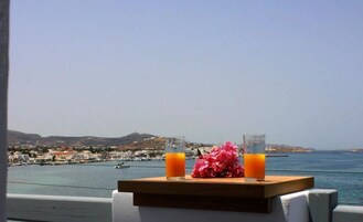 A table with two glasses of orange juice and a bouquet of pink flowers, overlooking a coastal town and the sea.