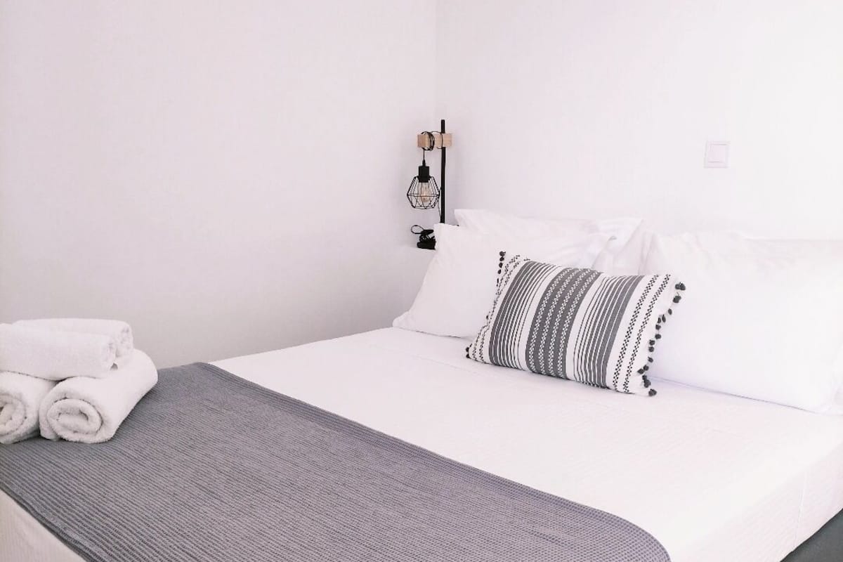A neatly made bed with white linens and a gray throw blanket, a small bedside table with a lamp, and a wall-mounted light fixture.