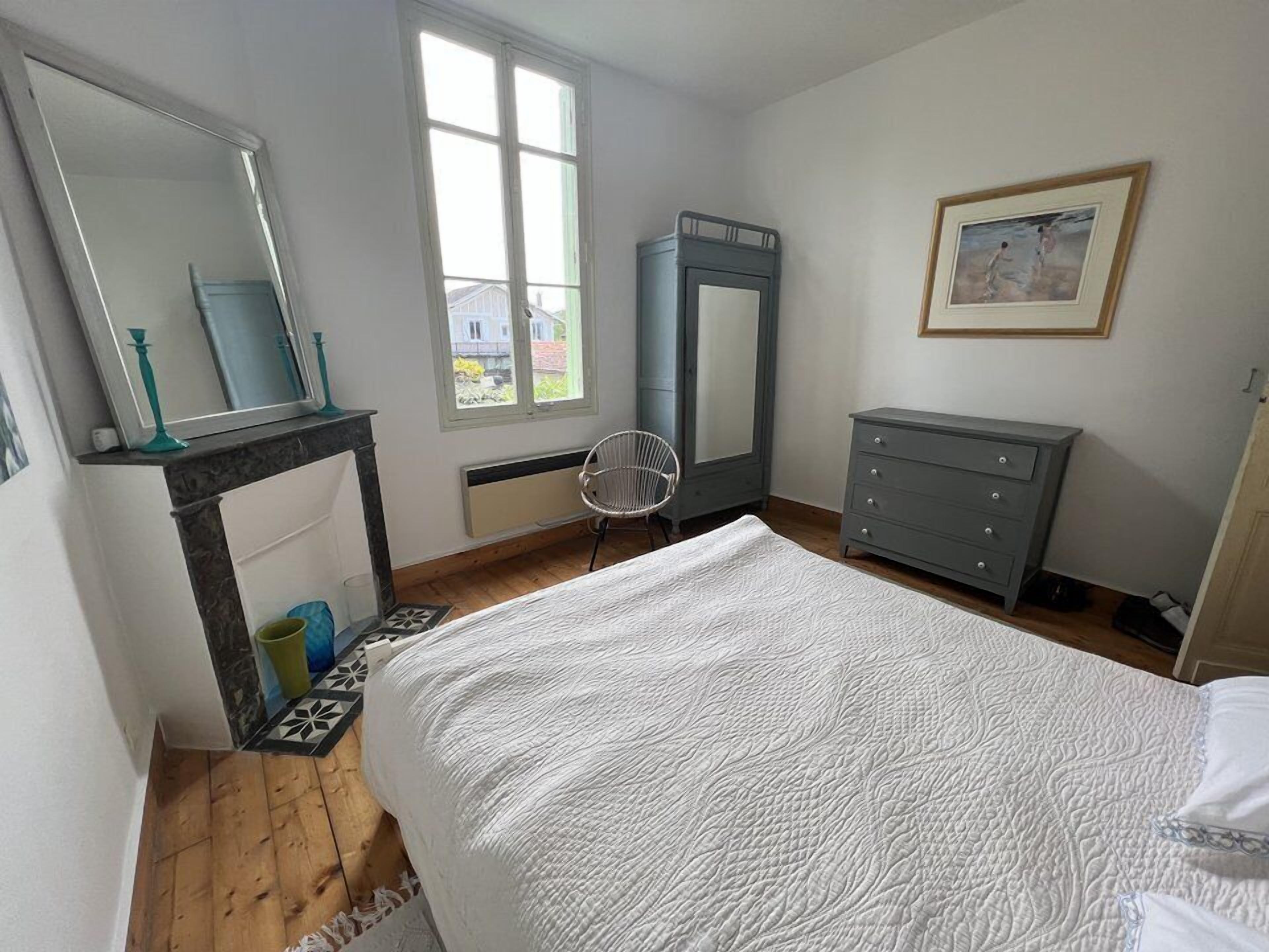 Une chambre à coucher avec un grand lit, une commode, une chaise, un miroir et une fenêtre donnant sur les maisons à l’extérieur.