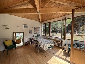 A room with a wooden ceiling, large windows, a table with chairs, a bookshelf, and a chair with a cushion. - Ref 546 - Real Soulacaise villa 2 steps from the city center (SOULAC SUR MER)