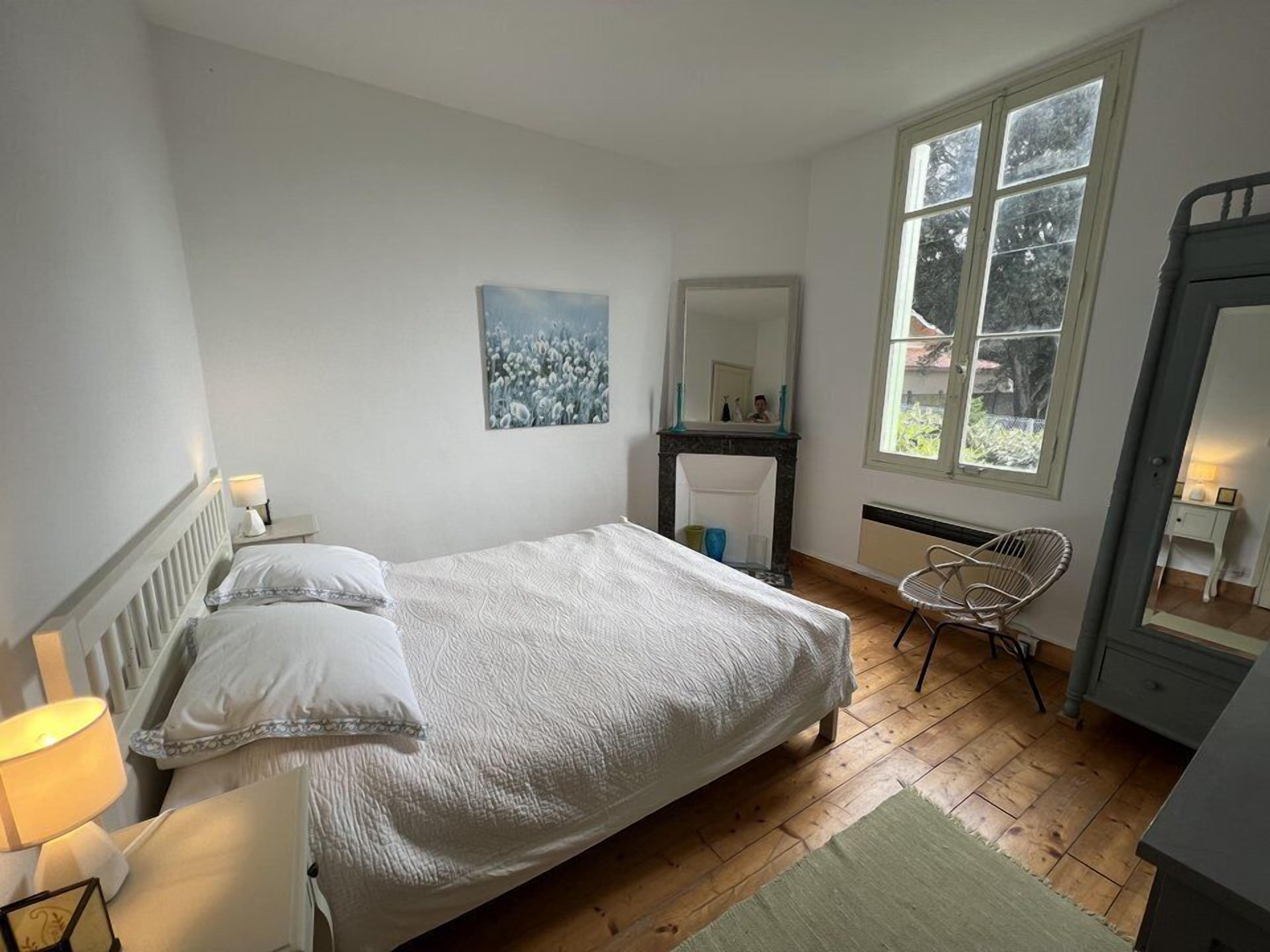 A bedroom with a bed, a nightstand, a chair, a fireplace, and a window with a view of trees.