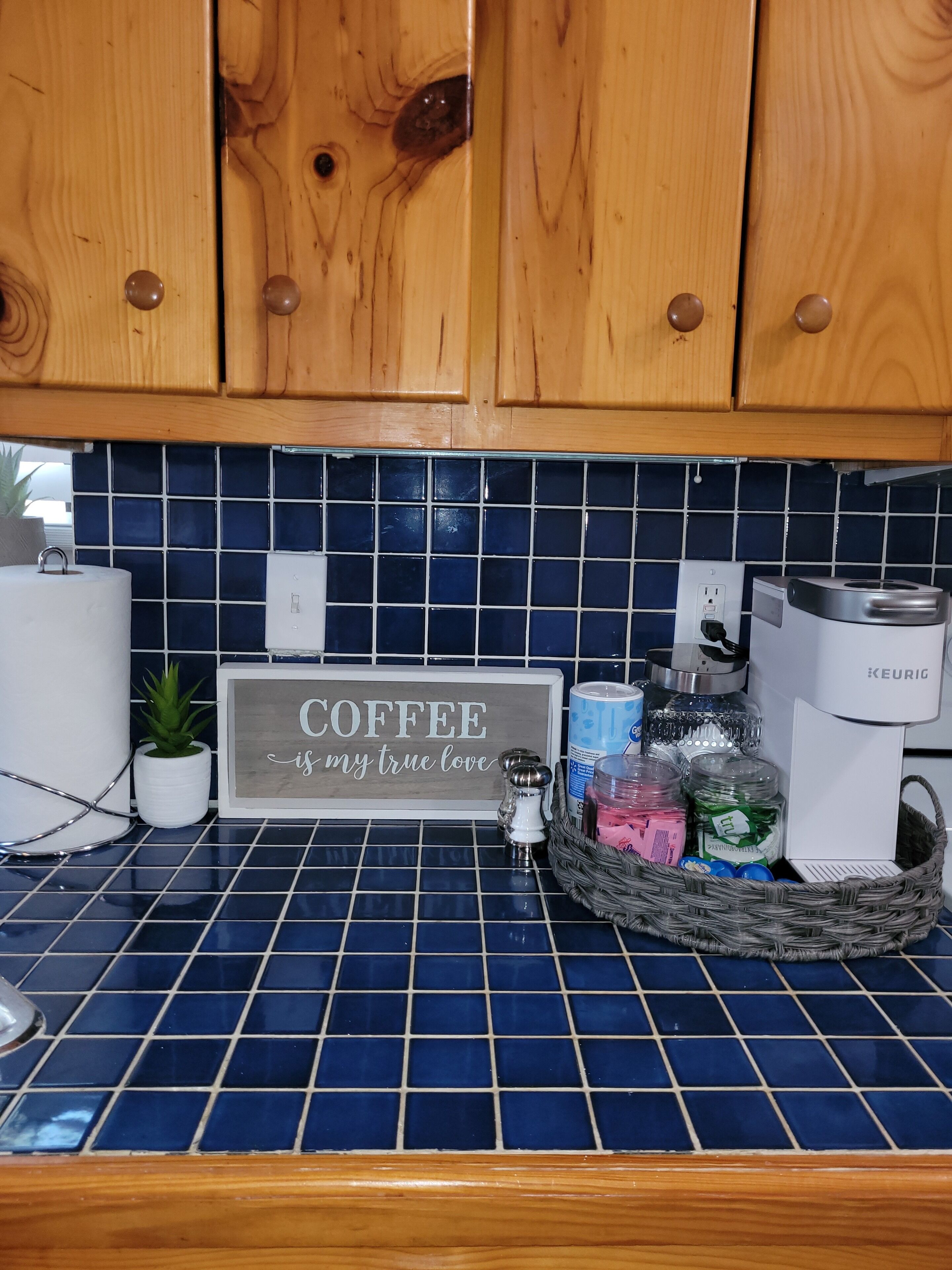 Cuina amb armaris de fusta, un frontissó blau de rajoles, un cartell de cafè i diversos objectes de cuina.