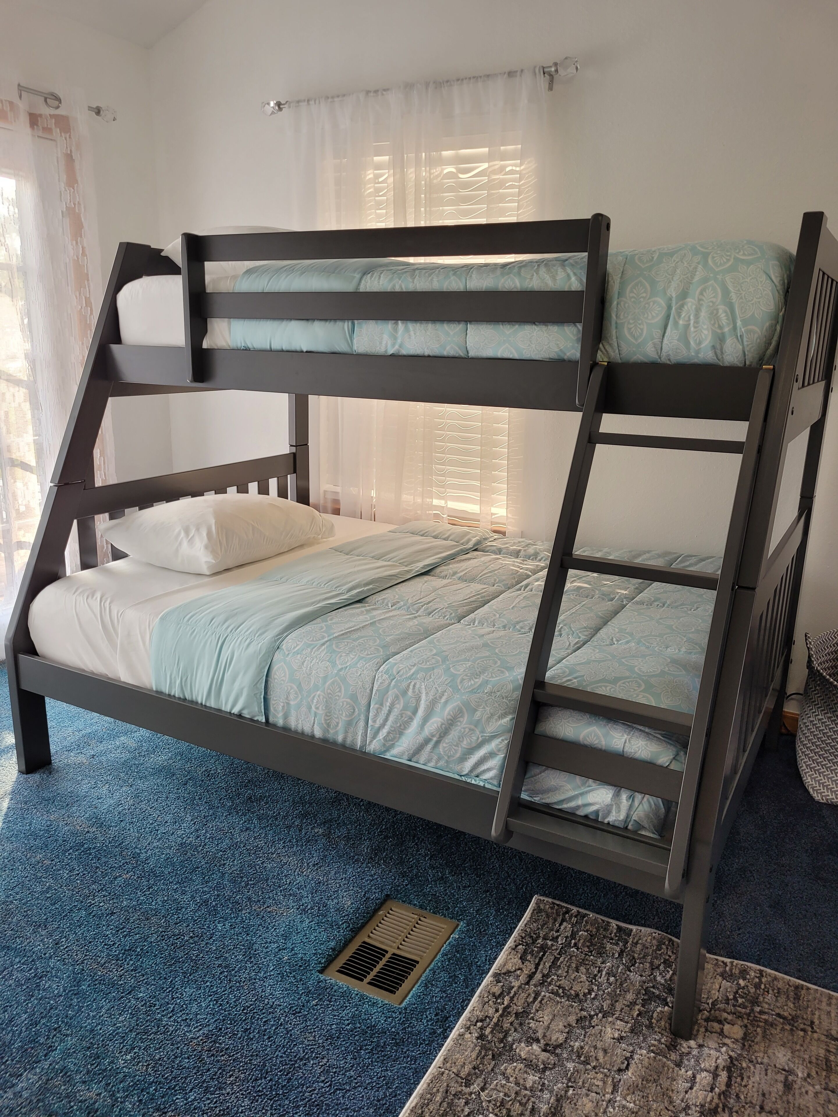 Una cama litera con una colcha y almohadas estampadas en azul, alfombra azul y una ventana con cortinas traslúcidas.