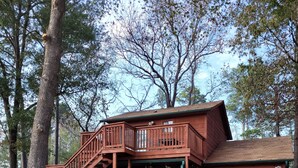A two-story wooden cabin with a balcony and a covered porch, surrounded by trees and a grassy area. - Modern meets Rustic Cabin on the Lake (Livingston)