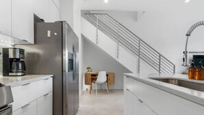 Une cuisine moderne dotée d’appareils électroménagers en acier inoxydable, un coin repas avec une table en bois et des chaises blanches, et un escalier avec une rampe en métal.