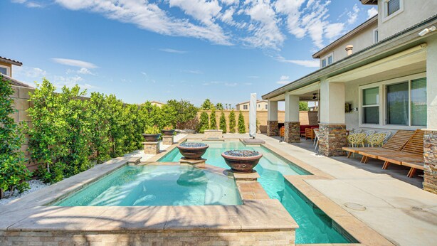 A swimming pool with two large stone planters, a covered patio area with outdoor furniture, and a view of neighboring houses. - Spacious Indio Home: Pool, Grill, & Fire Pits (Indio)