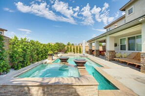 A swimming pool with two large stone planters, a covered patio area with outdoor furniture, and a view of neighboring houses.