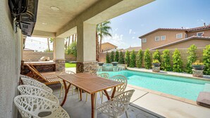 An outdoor patio with a pool, wooden table, white chairs, and a stone barbecue area. - Spacious Indio Home: Pool, Grill, & Fire Pits (Indio)