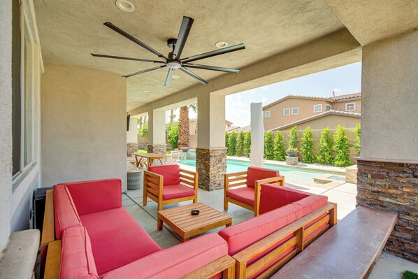 A covered outdoor patio with red cushioned seating, a ceiling fan, and a view of a swimming pool and residential buildings in the background.