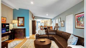 A living room with a brown leather sofa, a coffee table, a TV stand with streaming service logos, and a kitchen in the background.