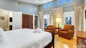 Une chambre d’hôtel avec un grand lit, deux fauteuils en cuir, une petite table avec une lampe et une vue sur la ville grâce à de grandes fenêtres.