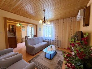 A living room with a sofa, a coffee table, and a plant. There is a kitchen area with cabinets and a window.
