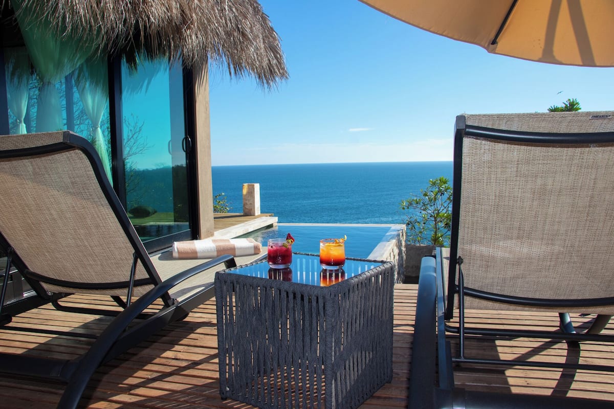 Un balcon avec deux chaises, une petite table et une boisson. La vue comprend une étendue d’eau et un ciel dégagé.