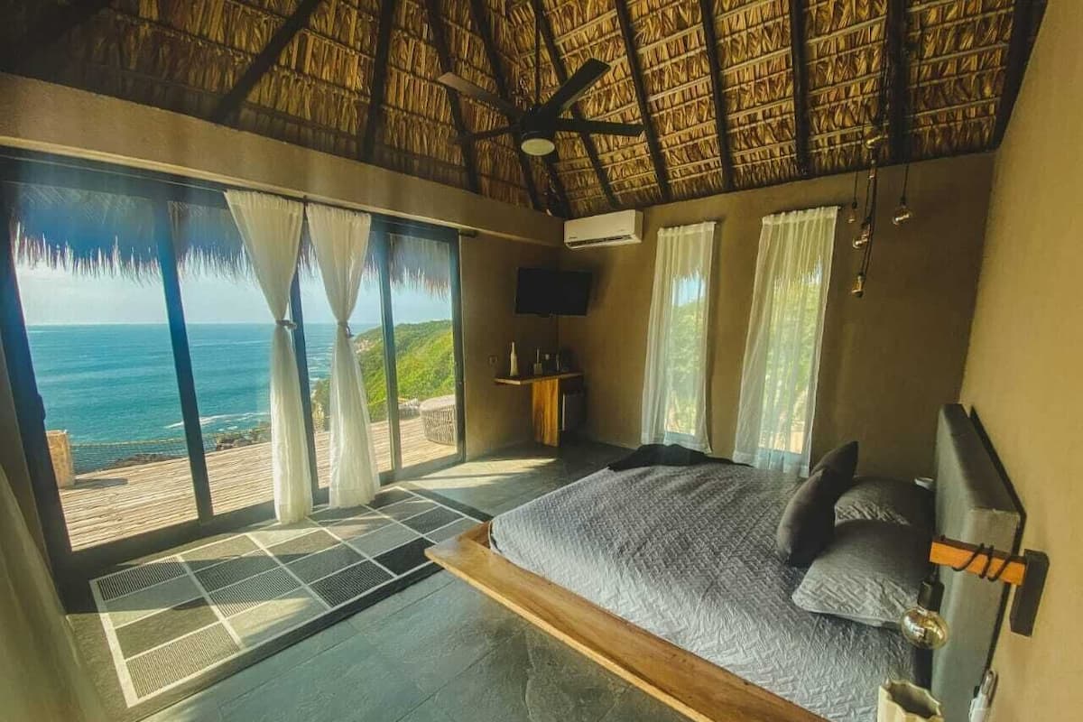 Une chambre avec un grand lit, un ventilateur de plafond, une télévision et une vue sur l’océan.