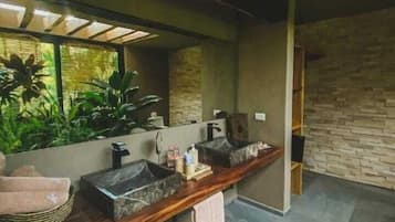 Relax in a serene space with a large mirror reflecting lush greenery, a double vanity with unique basins, and a cozy basket at your feet.