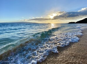 A beach with waves gently lapping onto the shore, a clear sky, and the sun setting in the distance.