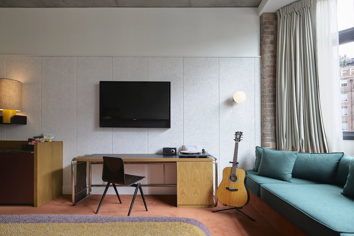 A modern living room with a flat-screen TV mounted on the wall, a wooden desk with a chair, a guitar, and a teal sofa.