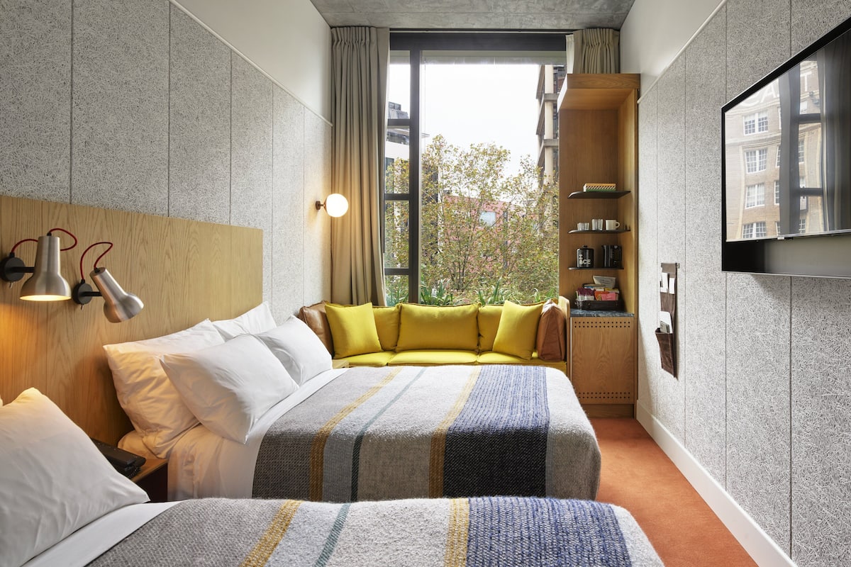 A hotel room with a bed, a sofa, a TV, and a window with a view of buildings and trees.