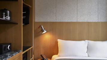 Habitación de hotel con un armario de madera, una cama con almohadas blancas, una mesita de noche con lámpara y un radio.