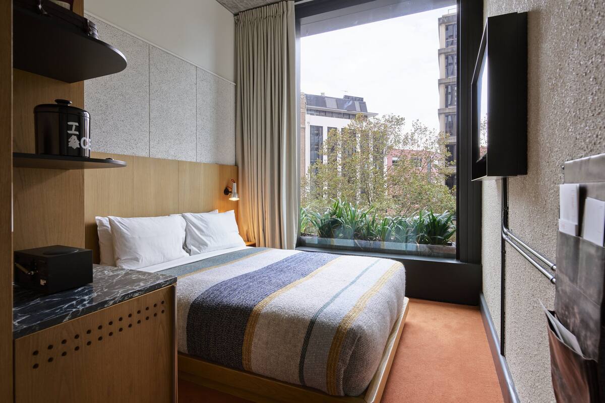 A hotel room with a bed, a desk, a TV, and a window with a view of buildings and trees.