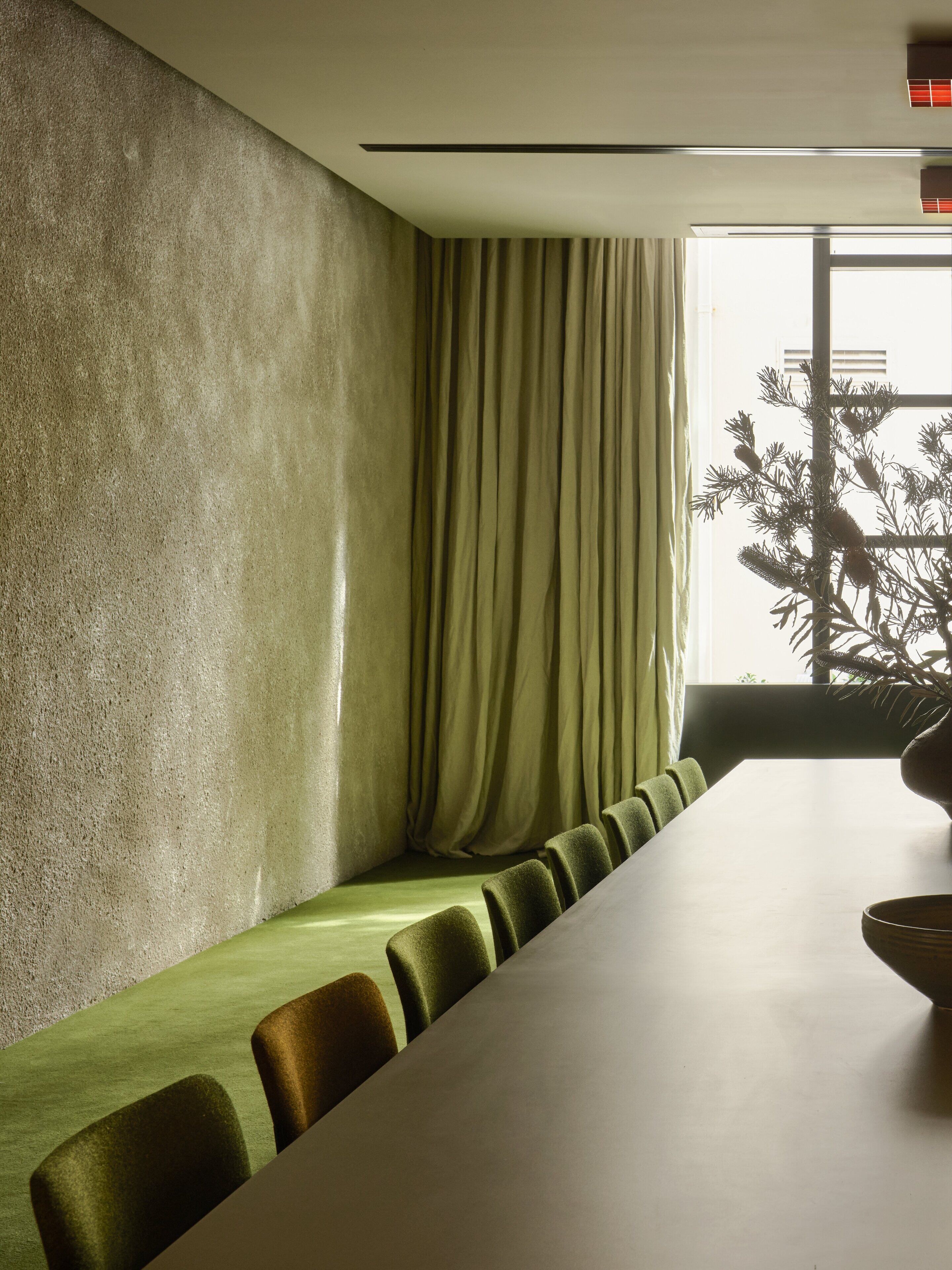 Een vergaderruimte met een lange tafel, groene stoelen, een structuurwand en een raam met gordijnen.