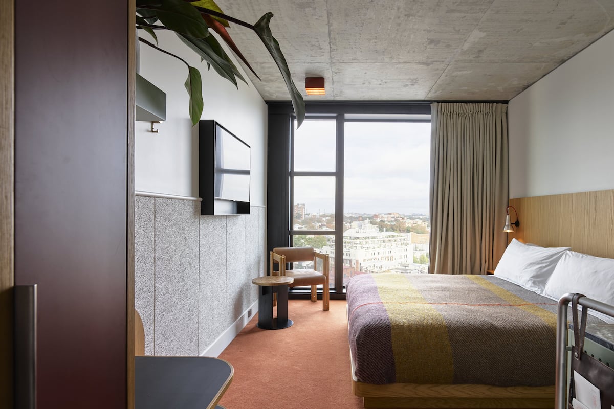 A hotel room with a large window, a bed, a chair, and a TV mounted on the wall.