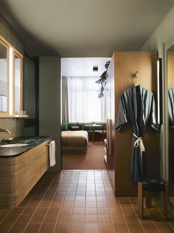 A compact hotel room with a wooden vanity, a bed, a small table, and a seating area with a view of the city. - Ace Hotel Sydney (Sydney)