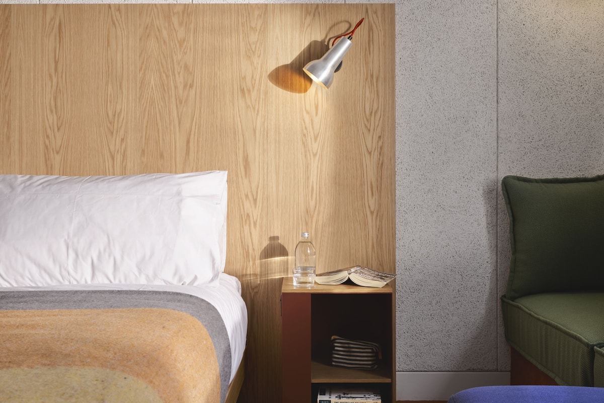A hotel room with a wooden headboard, a bedside table with a lamp, a green chair, and a bookshelf with books.