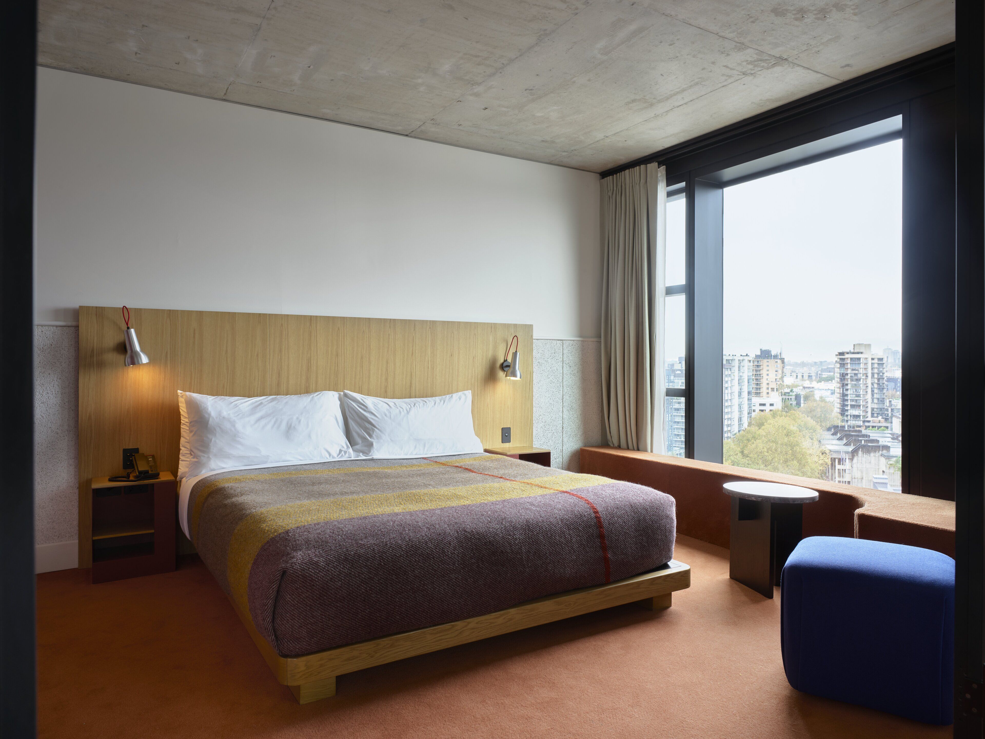 Una habitación de hotel moderna con una cama grande, cabecera de madera, lámparas de noche, una mesita y vistas a la ciudad a través de un ventanal.