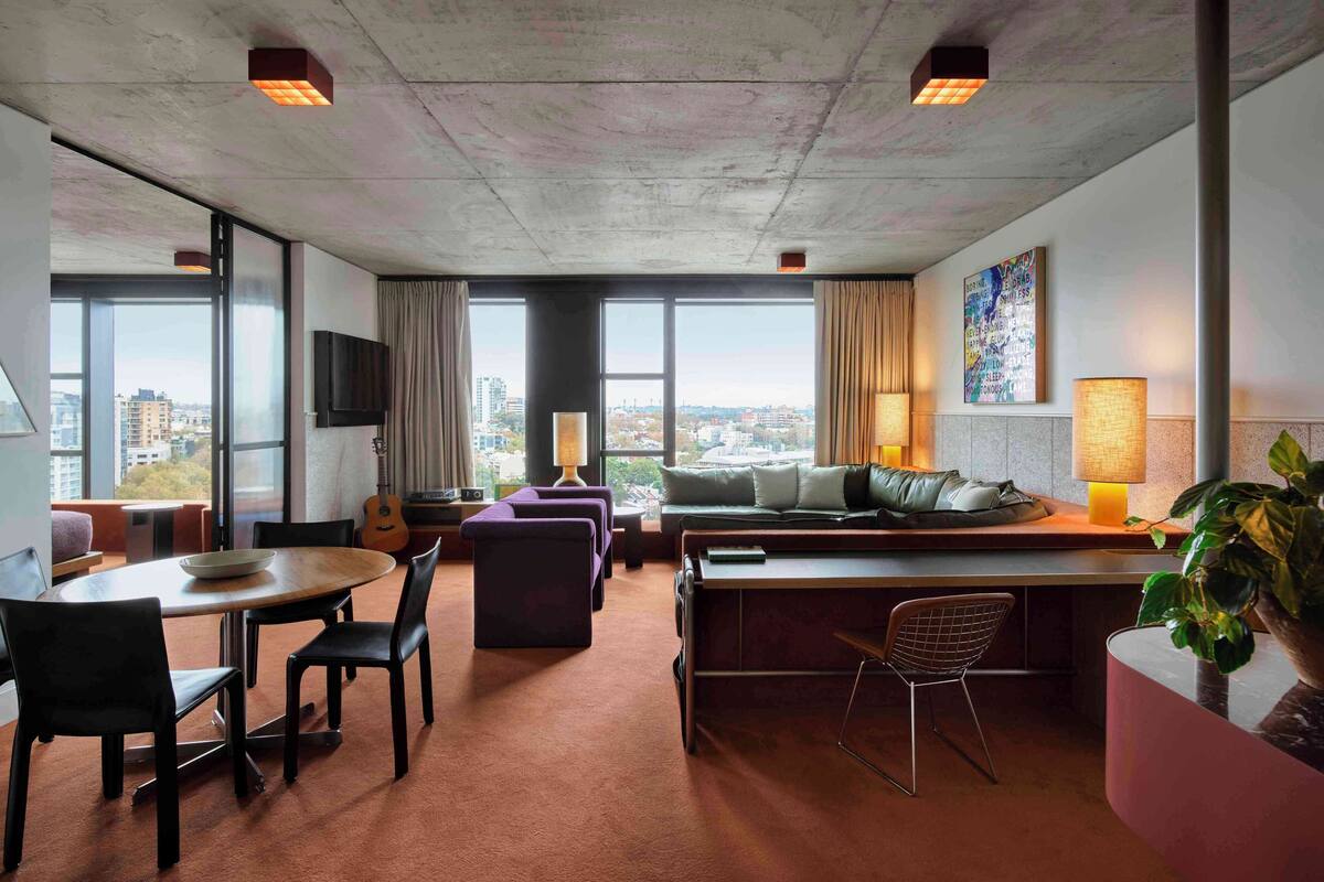 A modern hotel room with a large window, a dining area, a sofa, a TV, and a plant.