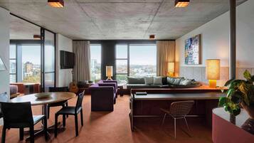 A modern hotel room with a large window, a dining area, a sofa, a TV, and a plant.