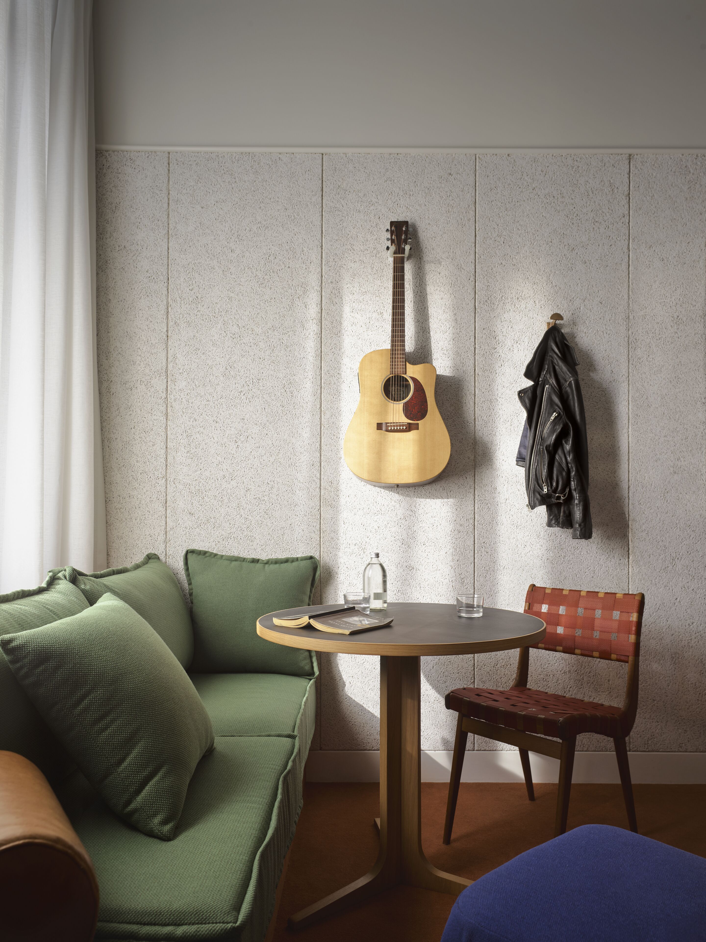 A room with a green sofa, a wooden table with a book and glasses, a guitar hanging on the wall, and a chair with a patterned backrest.