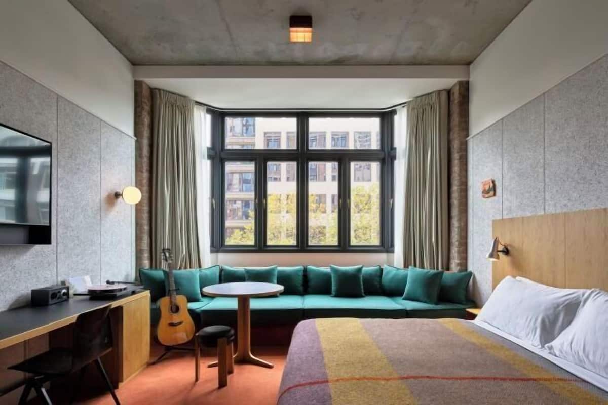A hotel room with a large window, a sofa bed, a desk, a chair, and a bed.