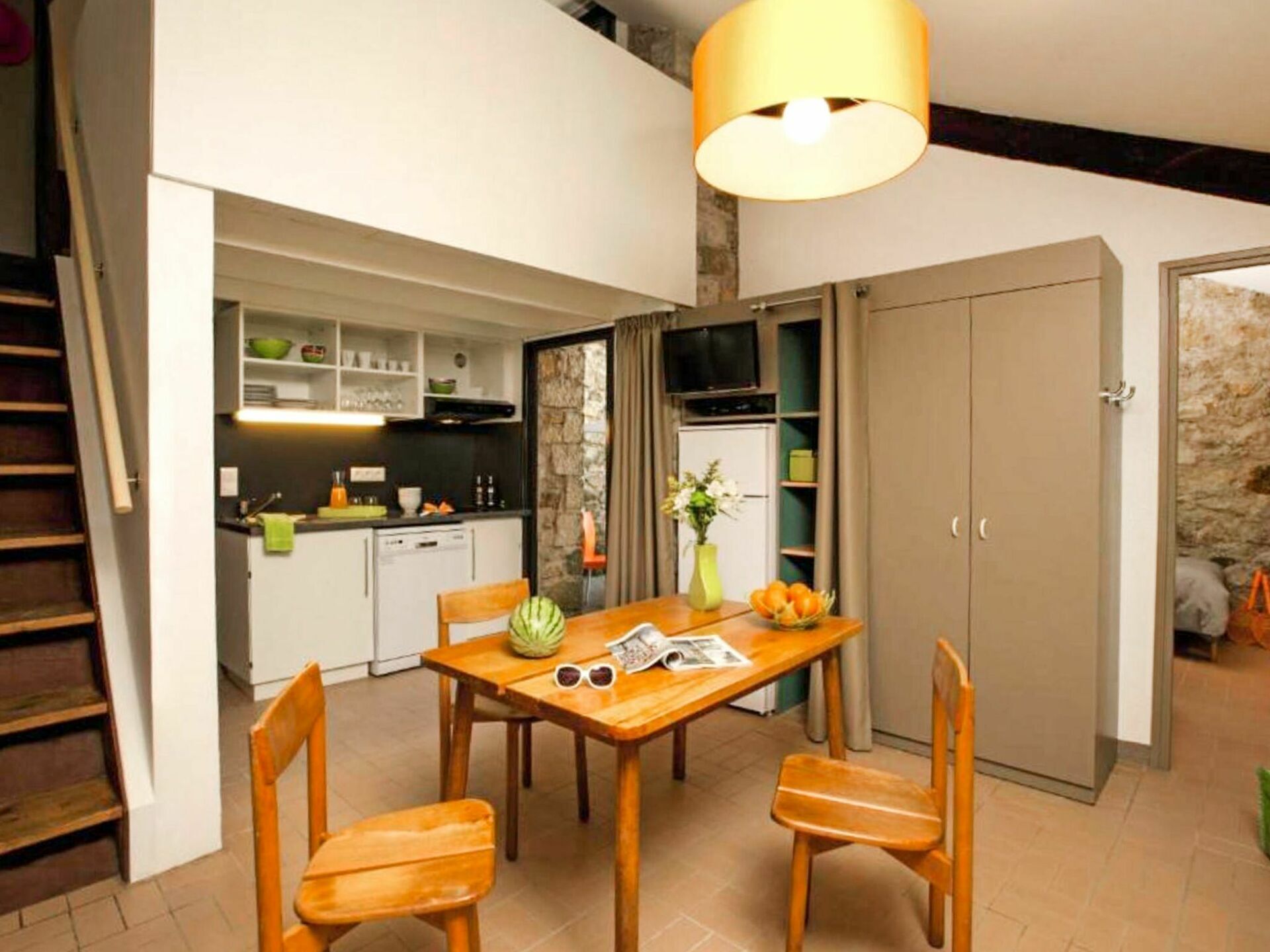 A compact kitchen with white cabinets, a dining table with four chairs, a wooden staircase, and a stone wall.