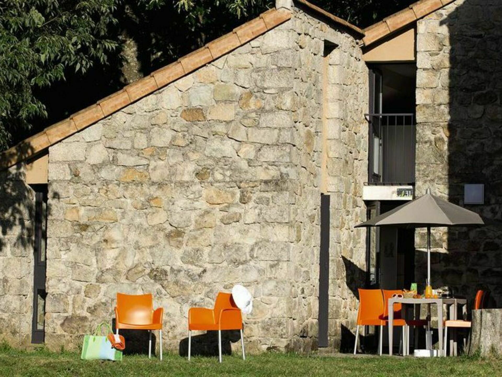 A stone building with a sloped roof, outdoor seating with orange chairs and a table, and a green bag on the ground.