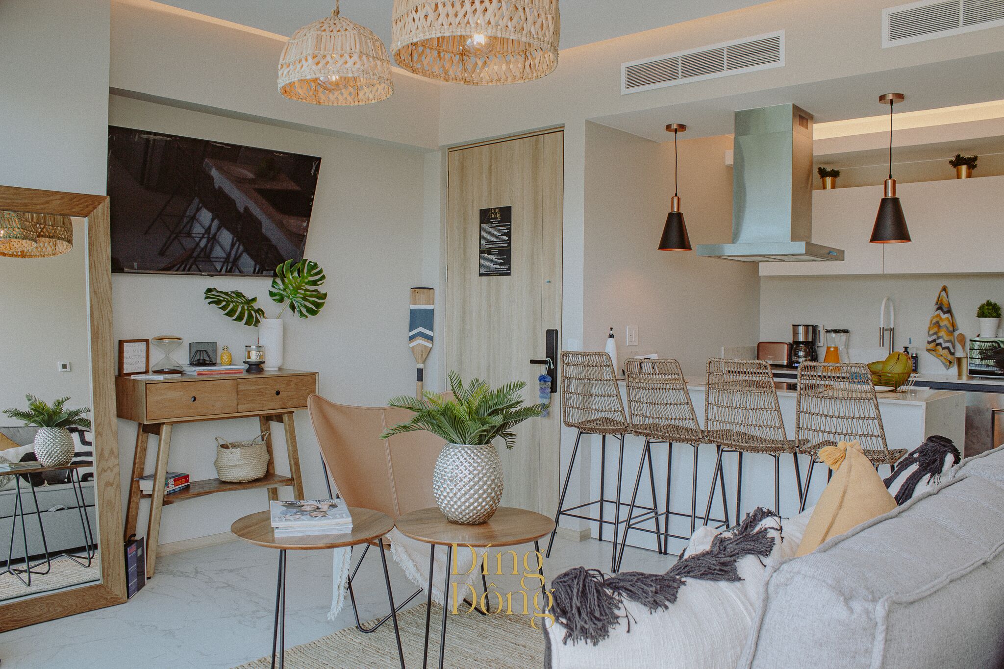 Un salon moderne avec un canapé, une table basse et des fauteuils, un coin repas avec des chaises en osier, et une cuisine équipée d’une cuisinière et de luminaires suspendus.