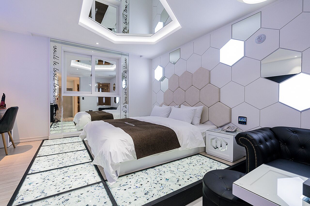Quarto de hotel moderno com uma cama grande, um sofá de couro preto, uma mesa de cabeceira branca e uma mesa de centro de vidro.