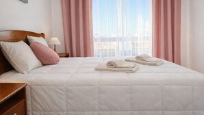 A neatly made bed with pillows, a bedside lamp, and rolled towels on a hotel room with a view through sheer curtains.