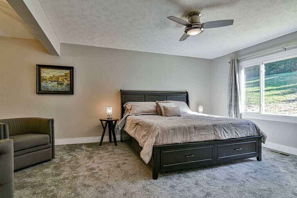 Een slaapkamer met een bed, een stoel, een nachtkastje, een plafondventilator en een raam met uitzicht op de omgeving.