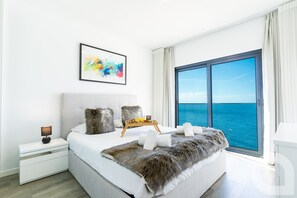 Ein modernes Schlafzimmer mit einem großen Bett, einem Nachttisch, einem gerahmten abstrakten Gemälde und Meerblick durch ein großes Fenster.