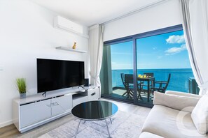Ein modernes Wohnzimmer mit Fernseher, einer Couch, einem Couchtisch und einem Balkon mit Meerblick.