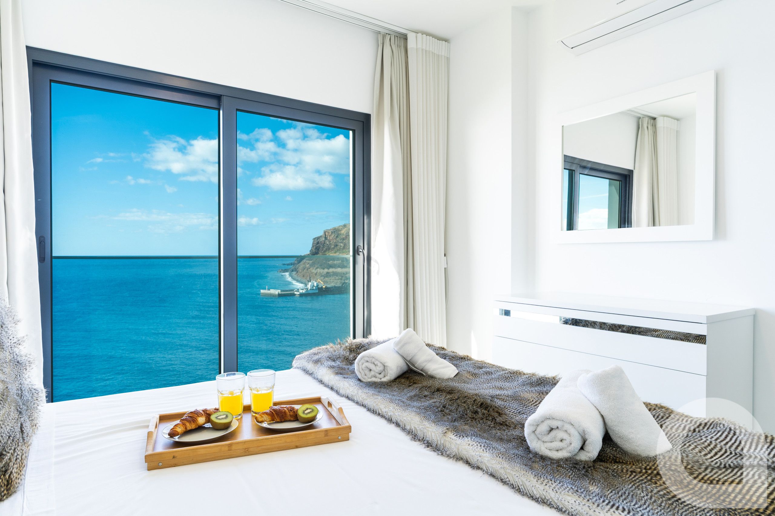 A hotel room with a large window offering a view of the sea and cliffs, a bed with a fluffy rug, towels, and a tray with croissants, juice, and a kiwi.