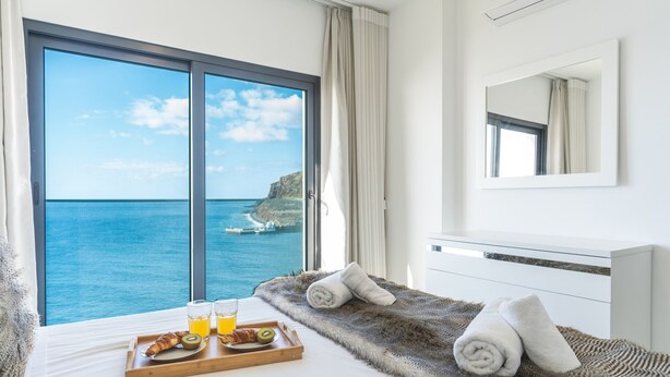 Une chambre d’hôtel dotée d’une grande fenêtre donnant sur la mer, d’un lit recouvert d’une couverture moelleuse et agrémenté de serviettes, ainsi que d’un plateau de petit-déjeuner comprenant des croissants, des kiwis et du jus d’orange.